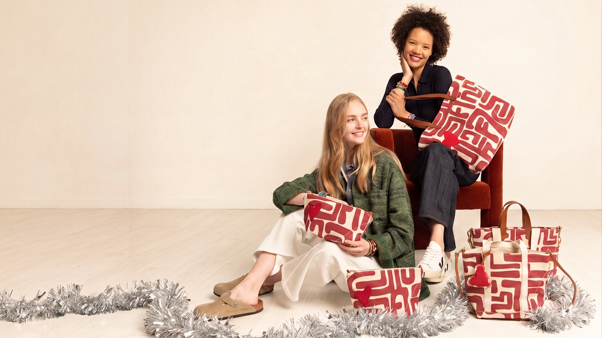 Two women pose with red and white patterned shopping bags and a matching tote bag in a minimalist studio setting with a beige wall and floor, decorated with silver tinsel.